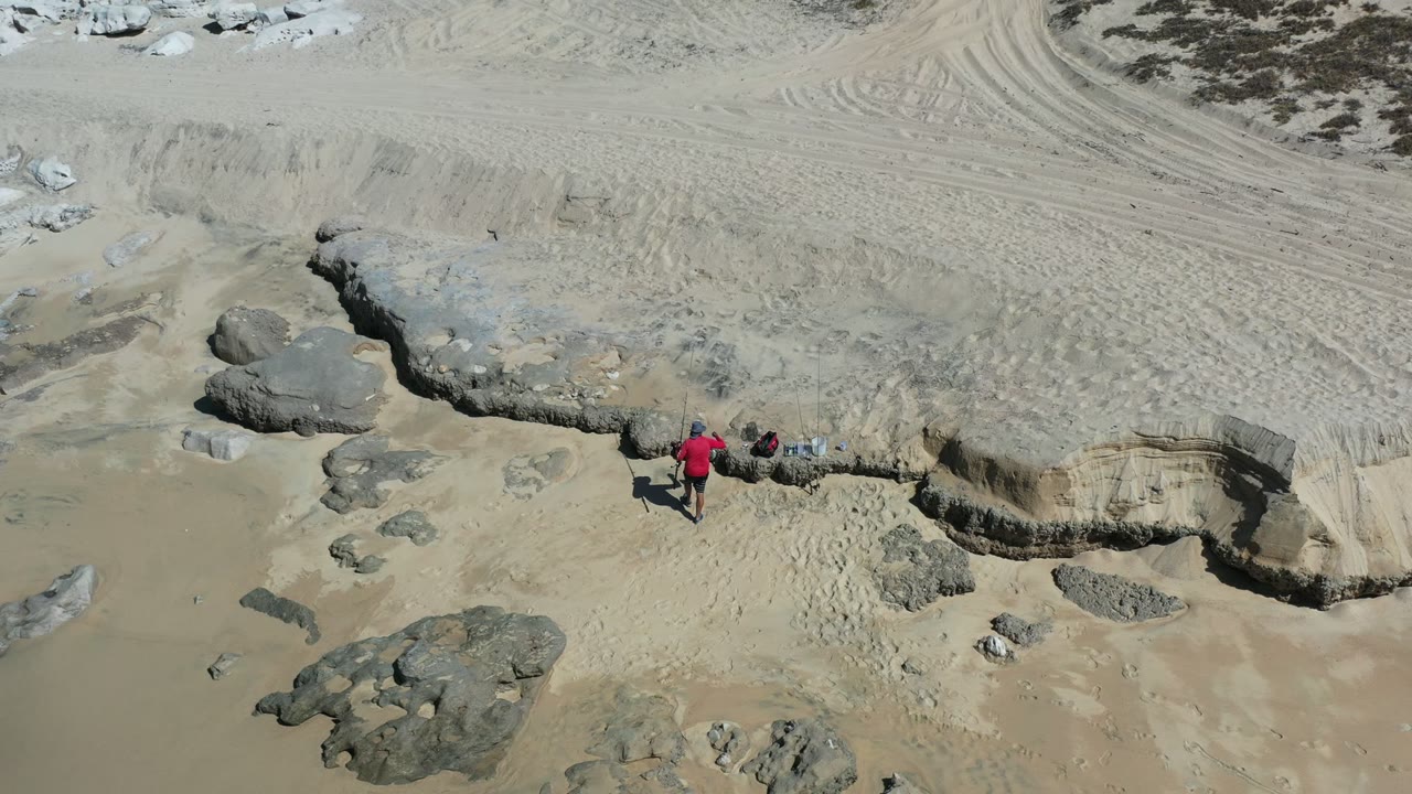 Overhead beach view with a person in a red shirt near eroded sand and rocks.