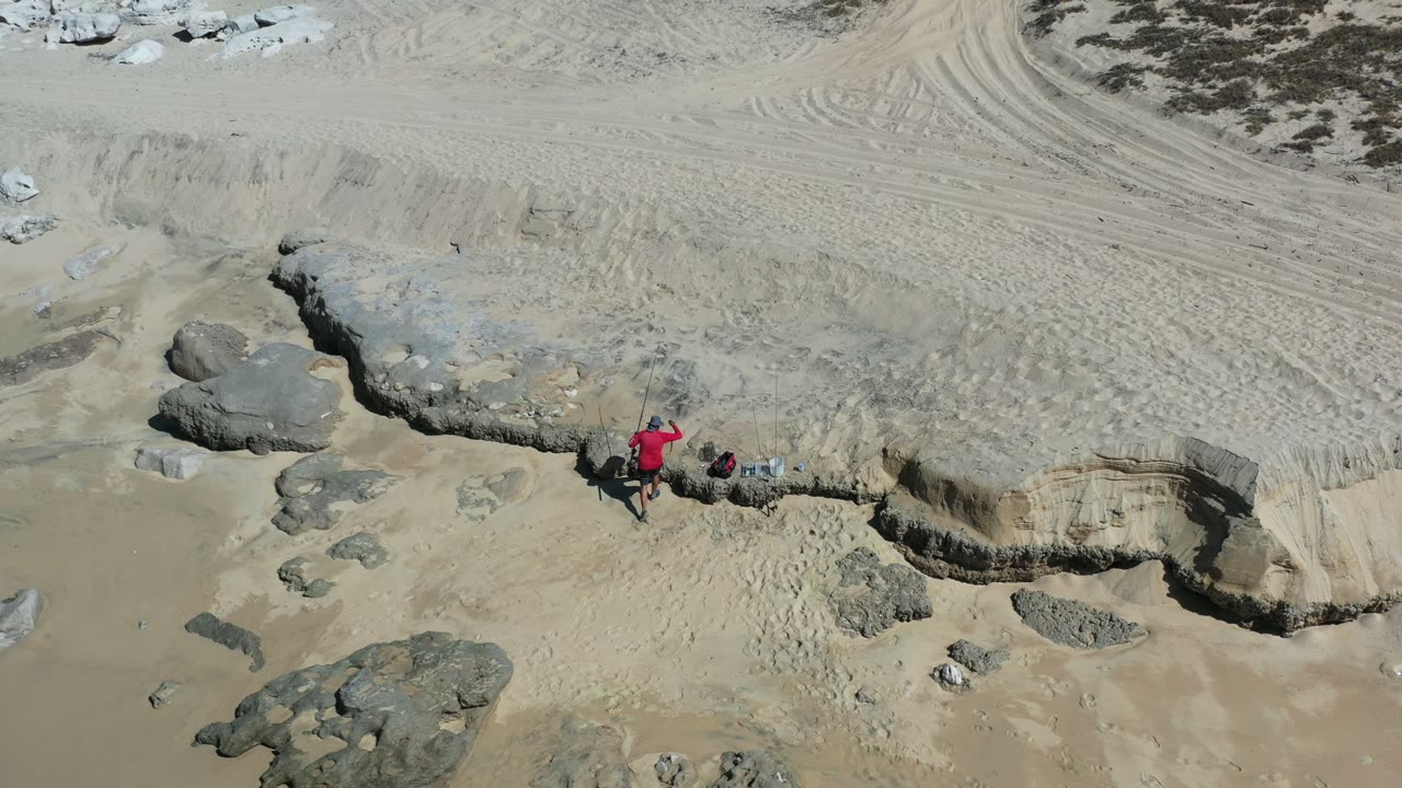 Aerial Los Cabos beach frame with the red-shirted person closer to small gear on the sand, beside layered erosion, rocks, wet sand, and faint surf wash.