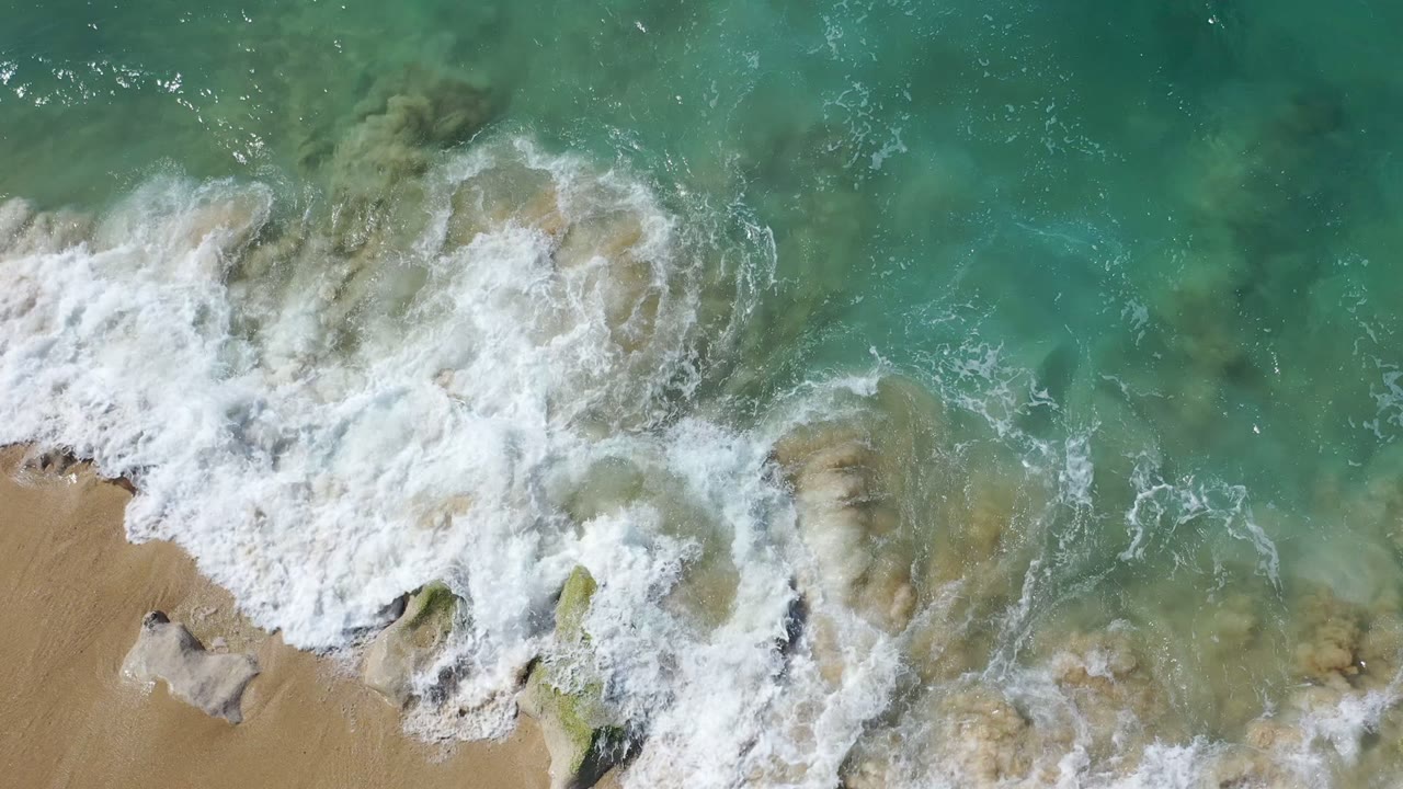 Los Cabos shoreline seen from above as a wider sheet of white foam spreads across shallow rocks and tan beach, with no visible people.