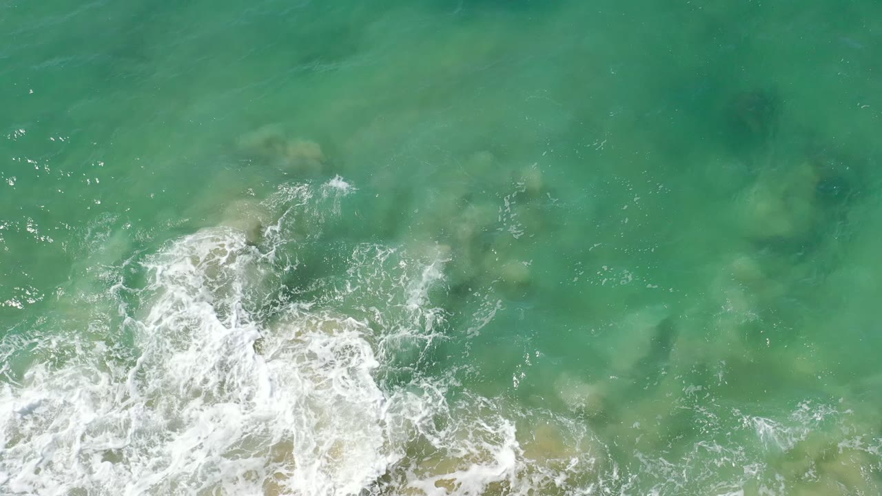 Overhead Los Cabos ocean frame with a larger white foam pattern spreading across translucent green water and no visible people.
