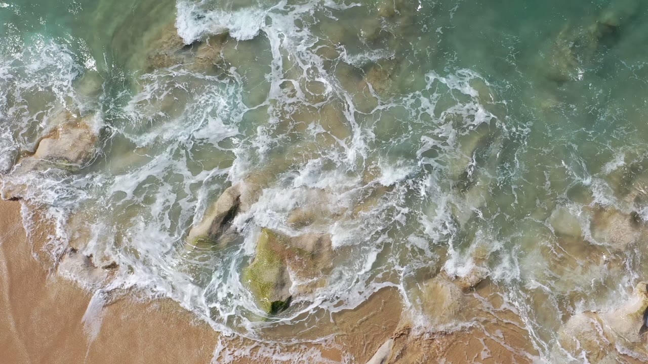 Aerial frame of pale green water and whitewash spreading over partly submerged rocks and tan beach in Los Cabos, with no visible people.