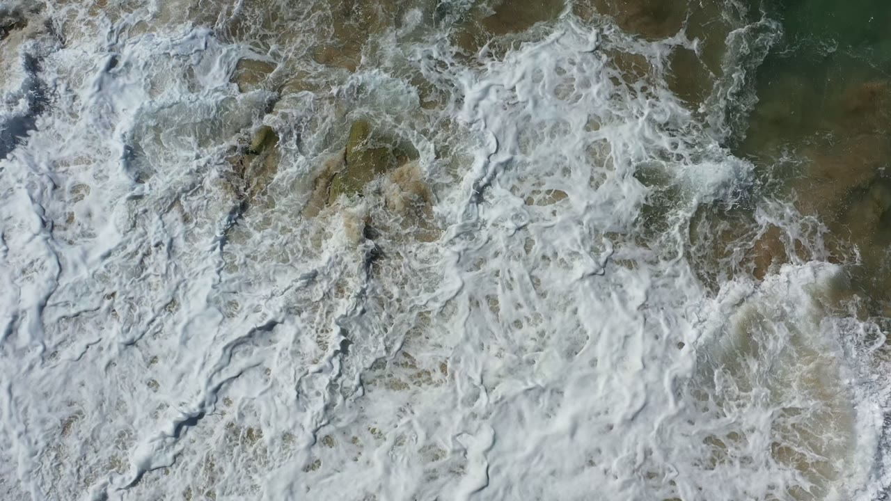 White wave foam forms streaks and patches over green-brown shallow water near wet sand in Los Cabos, Mexico, with no visible people.
