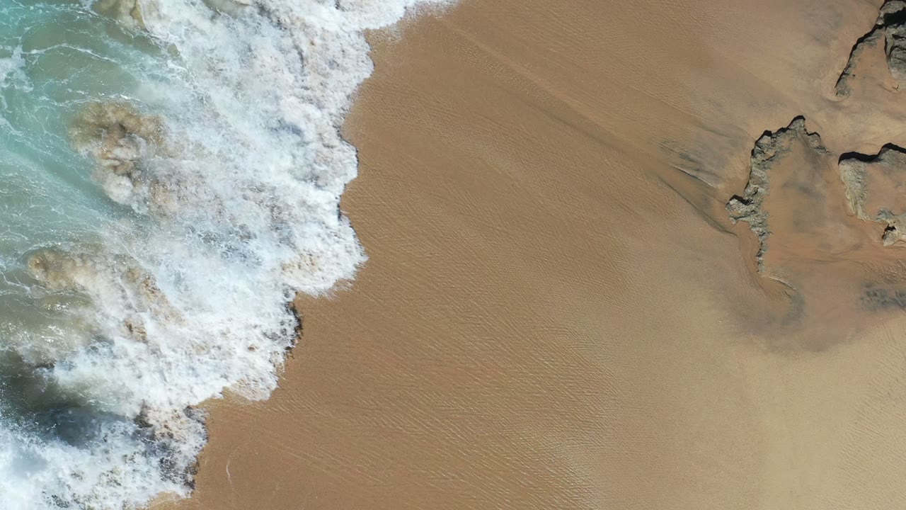 Overhead view in Los Cabos, Mexico of white surf entering from the upper left and spreading across tan sand near dark rock edges, with no visible people.