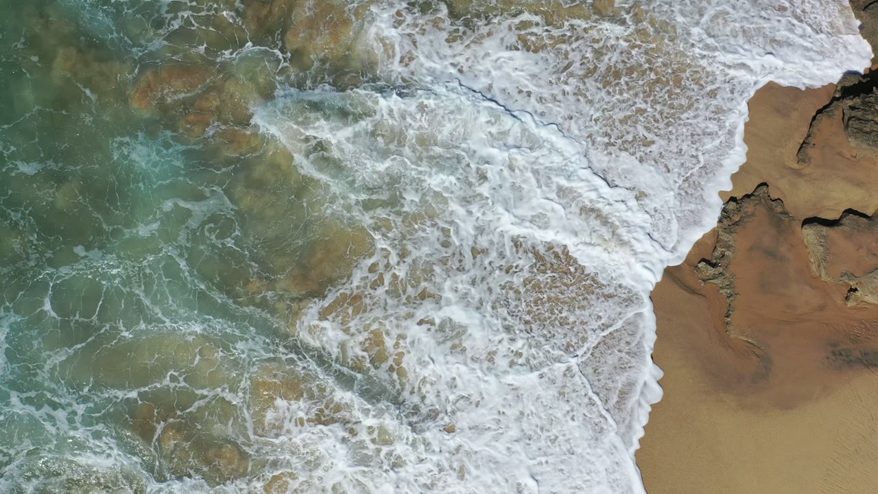 Overhead view of Los Cabos surf where dense whitewater fills the center and upper frame while tan sand and jagged dark rocks remain visible at the right, with no visible people.