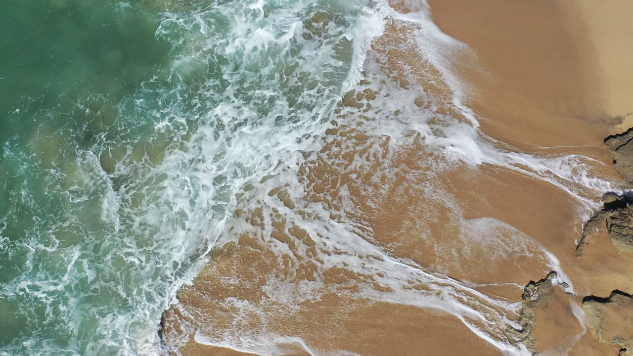 Aerial Los Cabos shoreline frame with white foam retreating across wet tan sand, green-blue water on the left, and dark rocks on the right, no visible people.