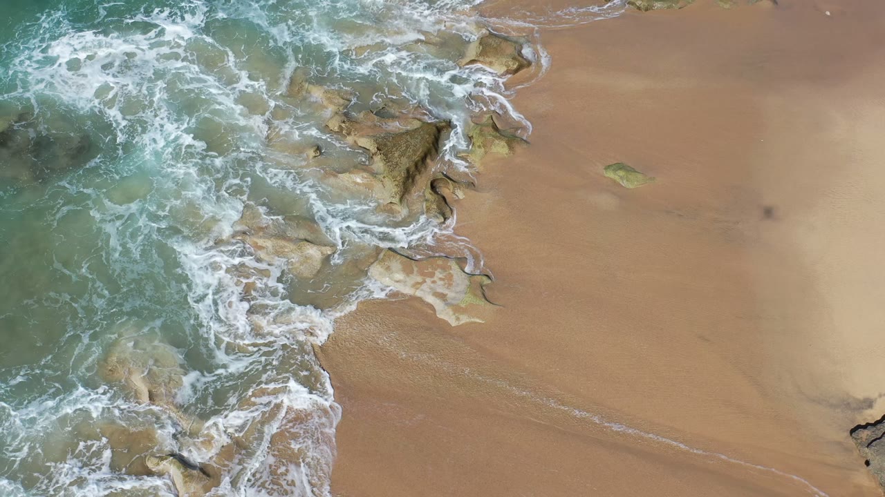 Overhead view in Los Cabos, Mexico of foamy surf curling around exposed beach rocks and tan sand, with no visible people.