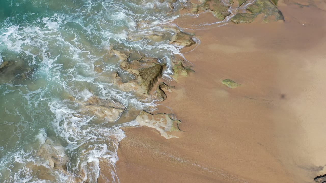 Aerial beach view in Los Cabos, Mexico with white foam tracing channels through shallow rocks and smooth tan sand, with no visible people.