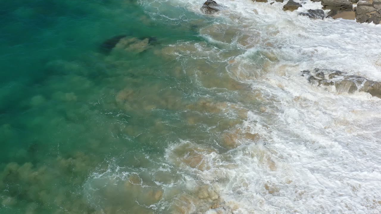 Los Cabos, Mexico shoreline from above, with green-blue water washing over submerged rocks and white foam entering from the right, no visible people.