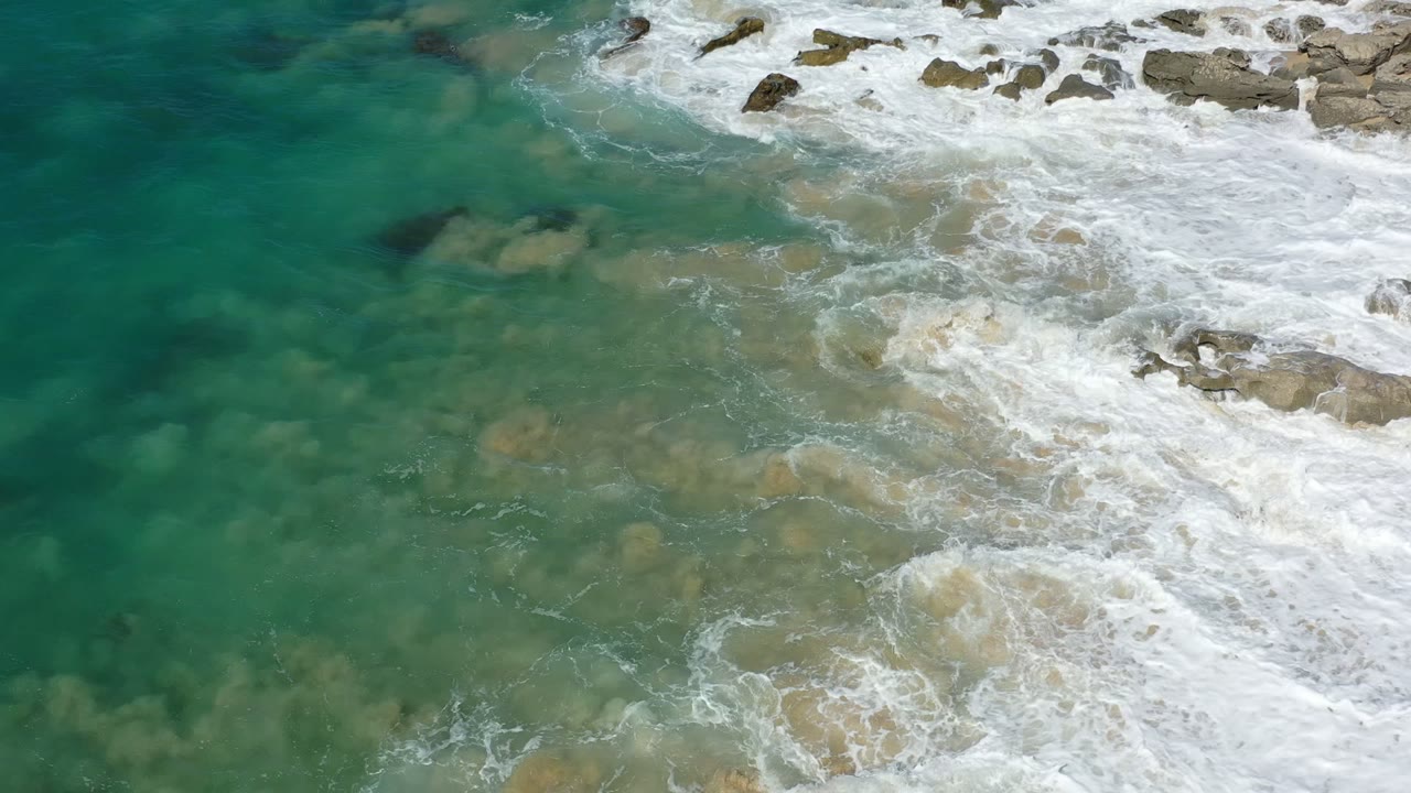 Los Cabos, Mexico rocky shoreline with clear green-blue water on the left and a wider patch of white surf curling across rocks on the right, no visible people.
