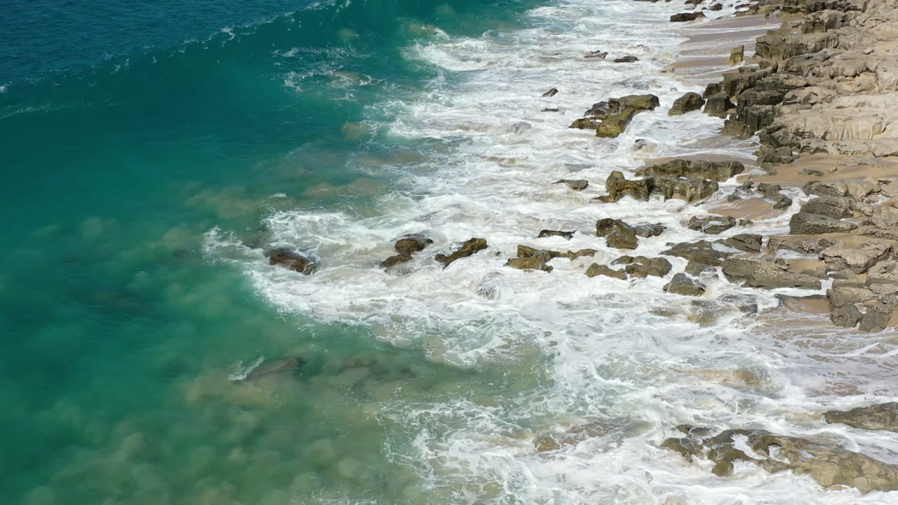 Aerial view of turquoise surf washing over rocks beside a sandy shore.