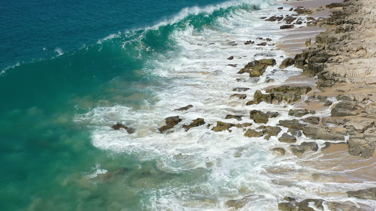 A green wave in Los Cabos, Mexico curls toward the rocky beach while earlier foam spreads across the shallow water, with no visible people.