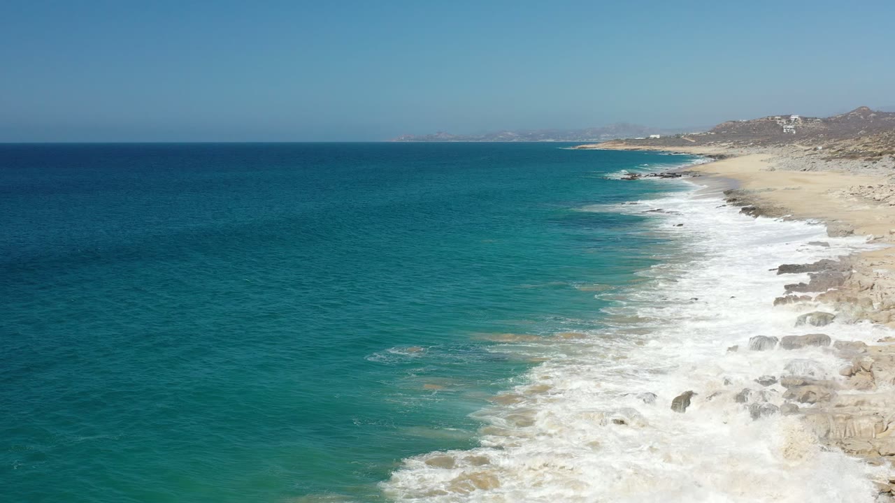 Elevated Los Cabos coastline view with turquoise water spreading toward white foam, tan rock shelves, pale beach, hazy hills, and no visible people.