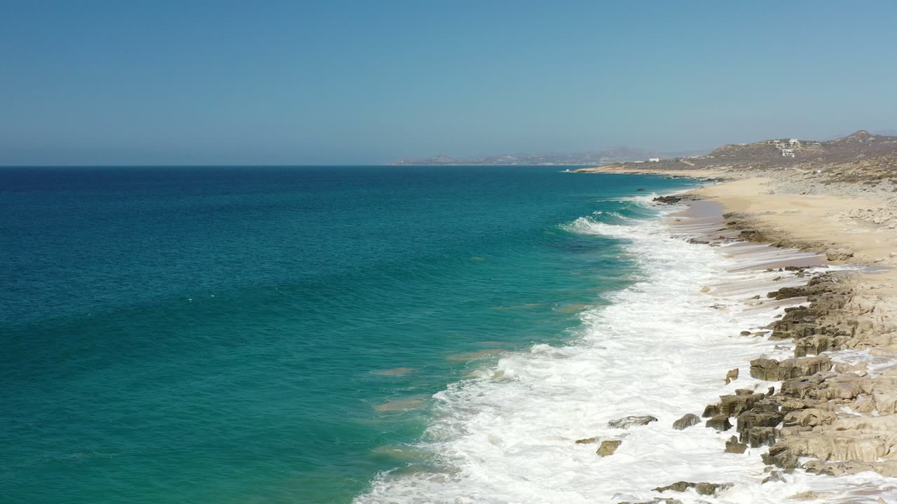 Aerial view of the Los Cabos coastline with turquoise water, white surf, tan sand, rocky shore, distant hills, and no visible people at the start of the clip.
