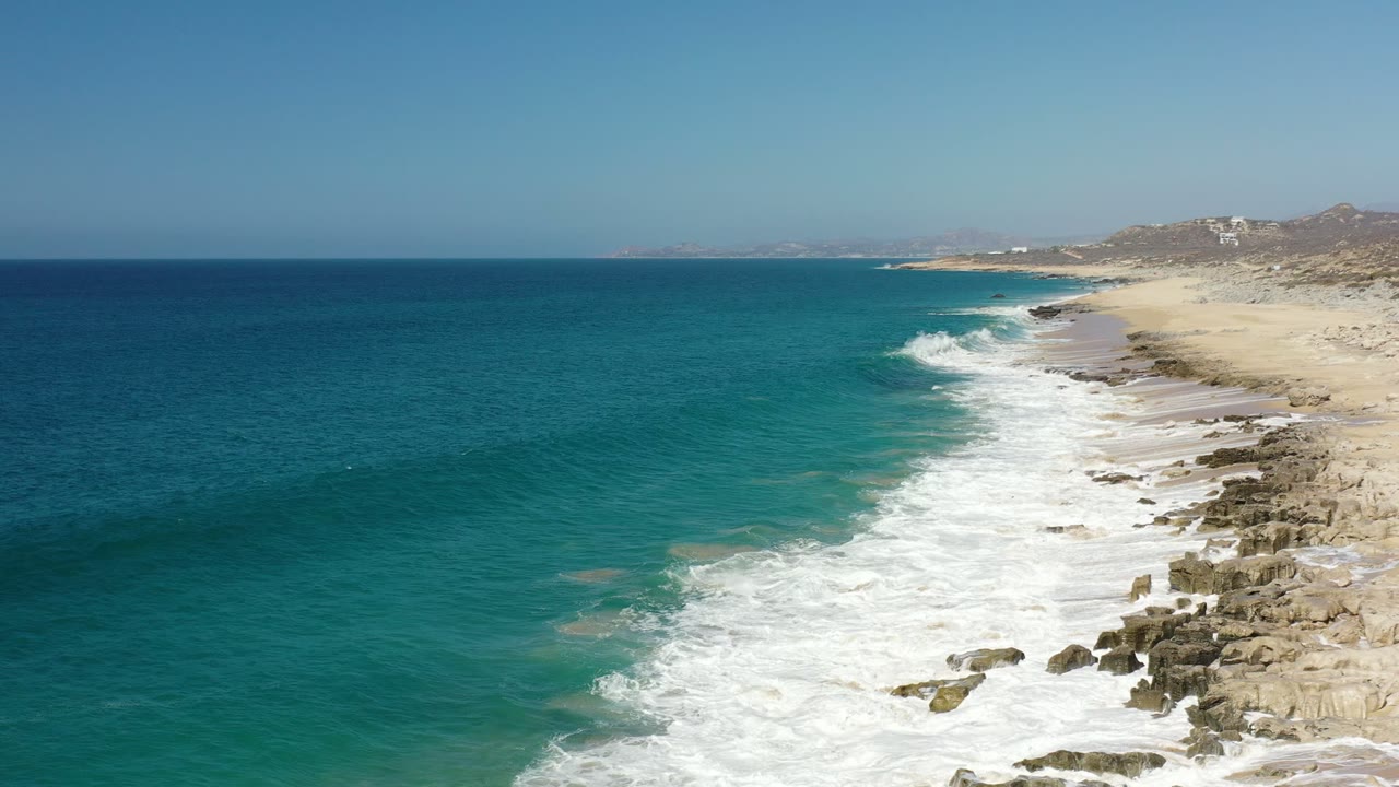 Los Cabos shoreline seen from above as a broad band of white water spreads along the rocks, with open blue-green ocean to the left and no visible people.