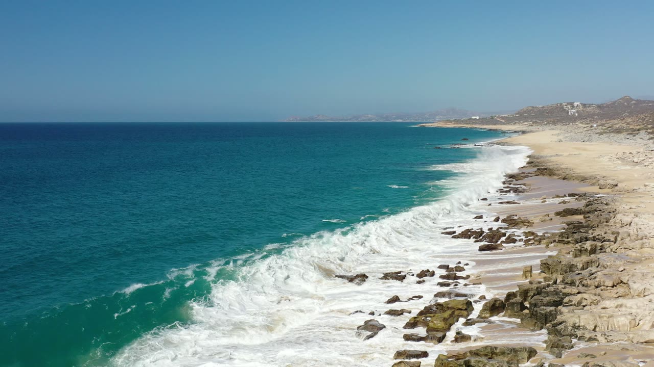 Aerial view in Los Cabos, Mexico with turquoise water at left, white surf crossing toward rocks and sand at right, distant hills on the horizon, and no visible people.