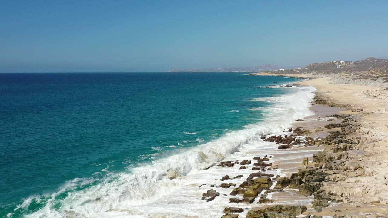 Aerial Los Cabos shoreline frame with a broader band of whitewater rolling over the rocky beach edge, turquoise sea to the left, tan sand to the right, and no visible people.