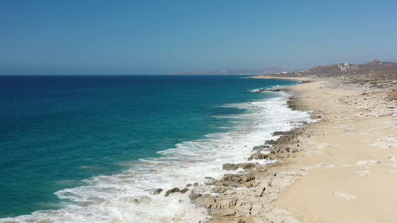 Wide aerial view of blue ocean surf meeting a rocky sandy Los Cabos coastline, with low hills and distant buildings on the horizon and no visible people.