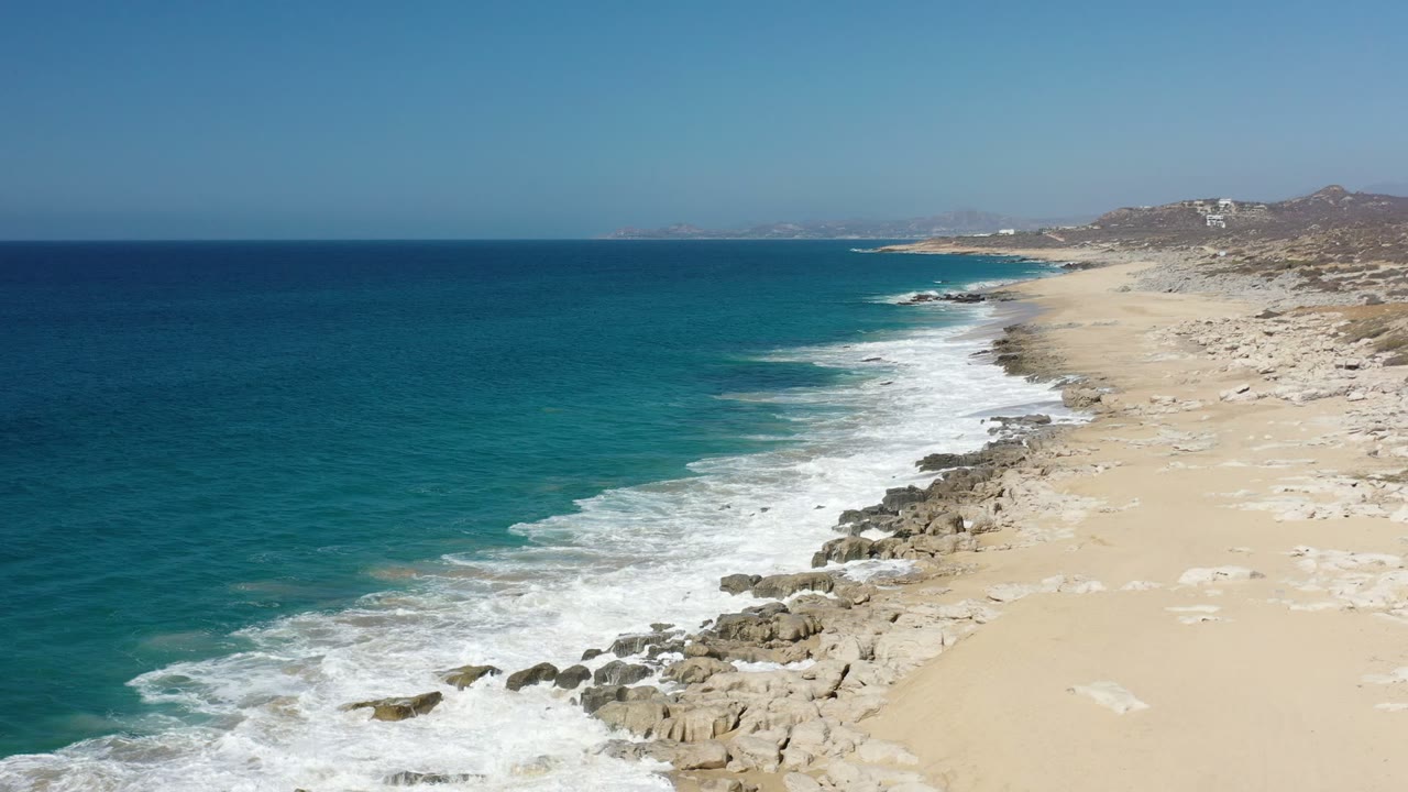 Aerial frame of white waves spreading across rocks and pale sand on the Los Cabos shore, with deep blue water to the left and no visible people.
