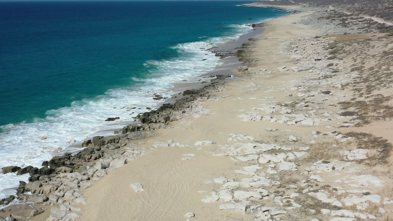 Aerial Los Cabos beach frame with foamy waves spreading farther along the rocky waterline, dry sand and low scrub visible inland, and no visible people.