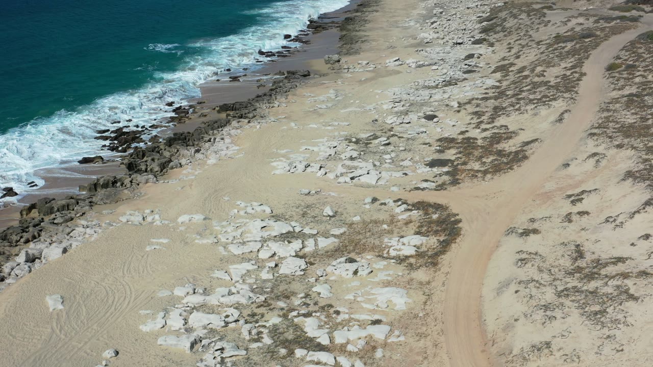 Aerial view of blue surf, rocky beach, coral-covered sand, and a sandy track.