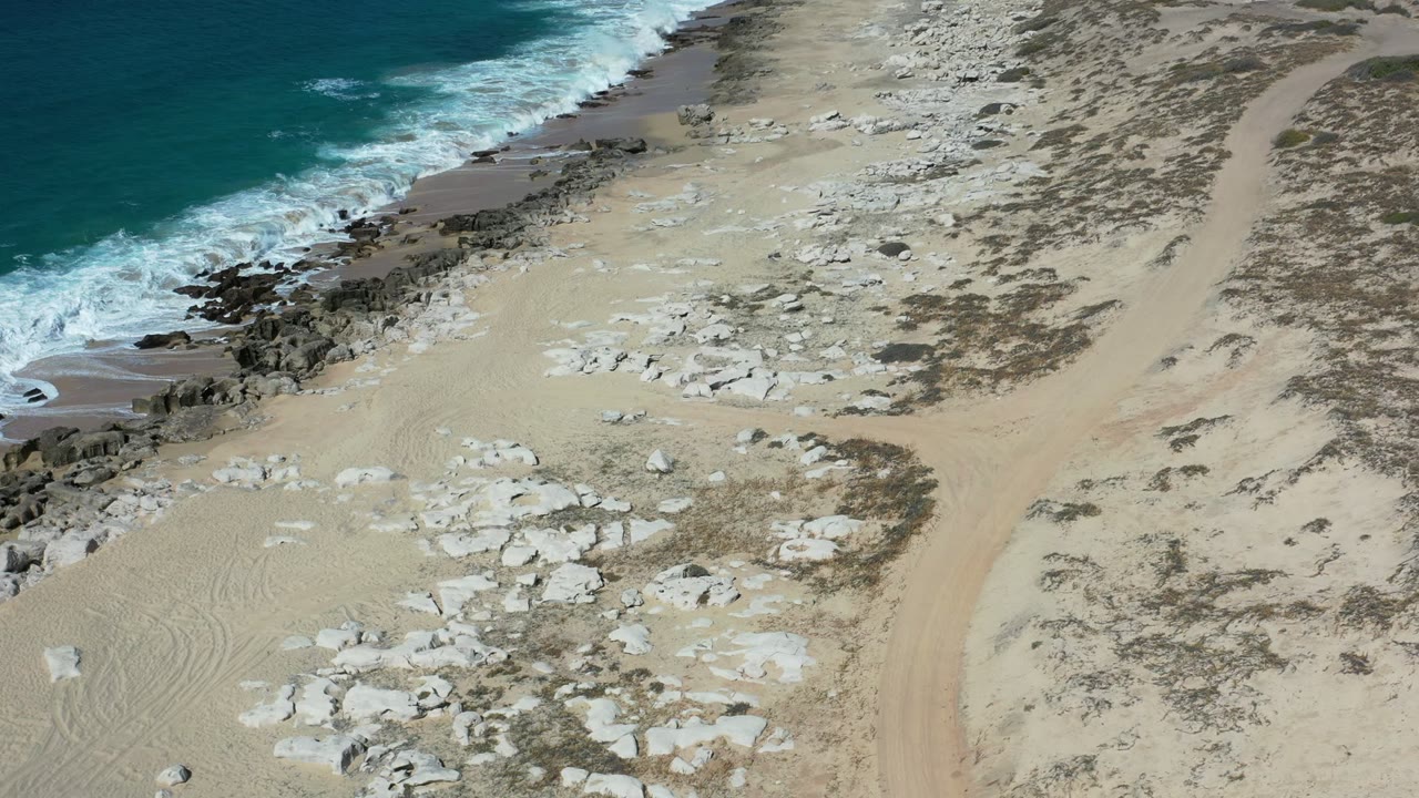 Aerial frame from Los Cabos, Mexico, showing surf breaking farther along the rocky edge, coral-like stones spread across the sand, a curving sandy track, and no visible people.