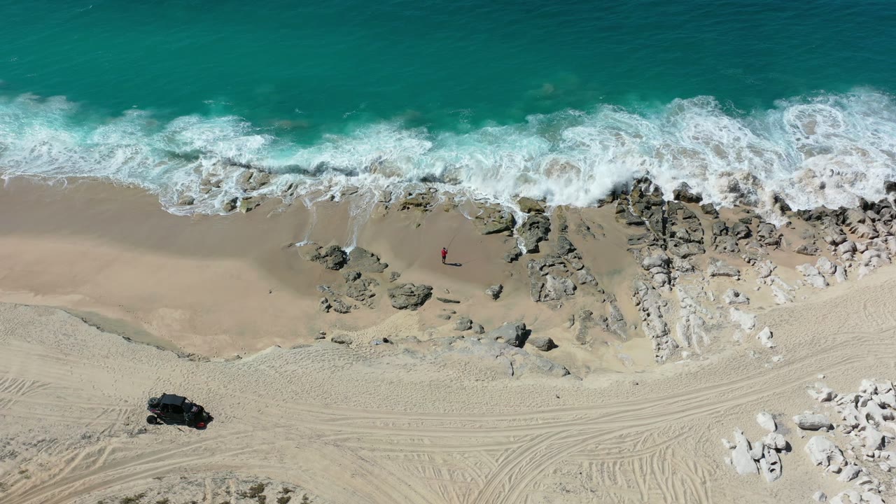 Overhead beach view with green water, white surf, rocks, sand tracks, and a dark vehicle-like shape.