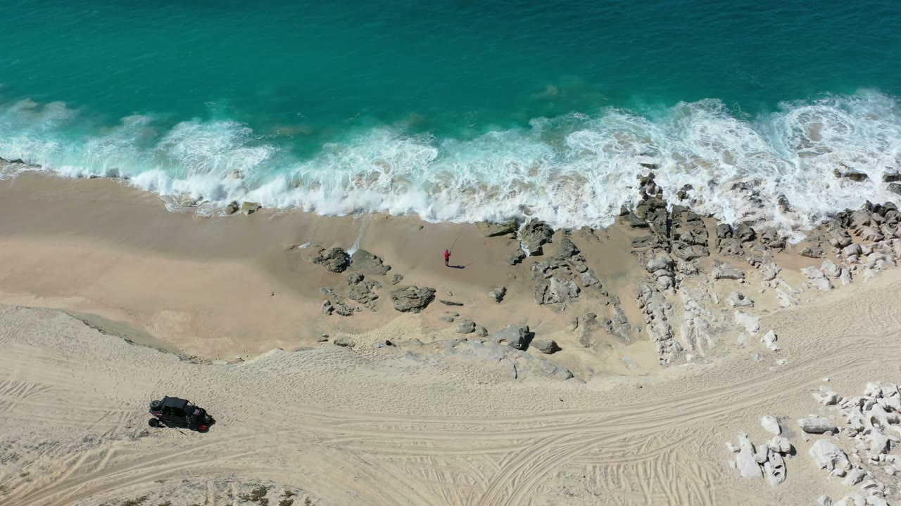 Overhead Los Cabos shoreline frame with a wider band of white foam across the center, rocky patches on the sand, tire tracks below, and no visible people.