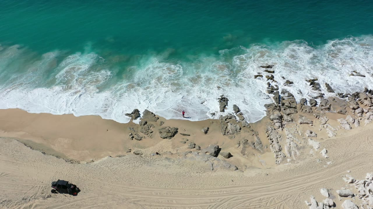 Overhead beach view with surf, rocks, sand, and tire tracks.