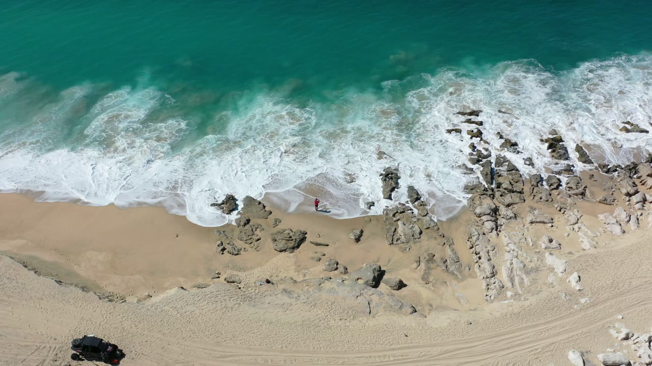 Aerial Los Cabos shoreline frame with foamy water spreading farther across the rock line and a broad dry sand band below.