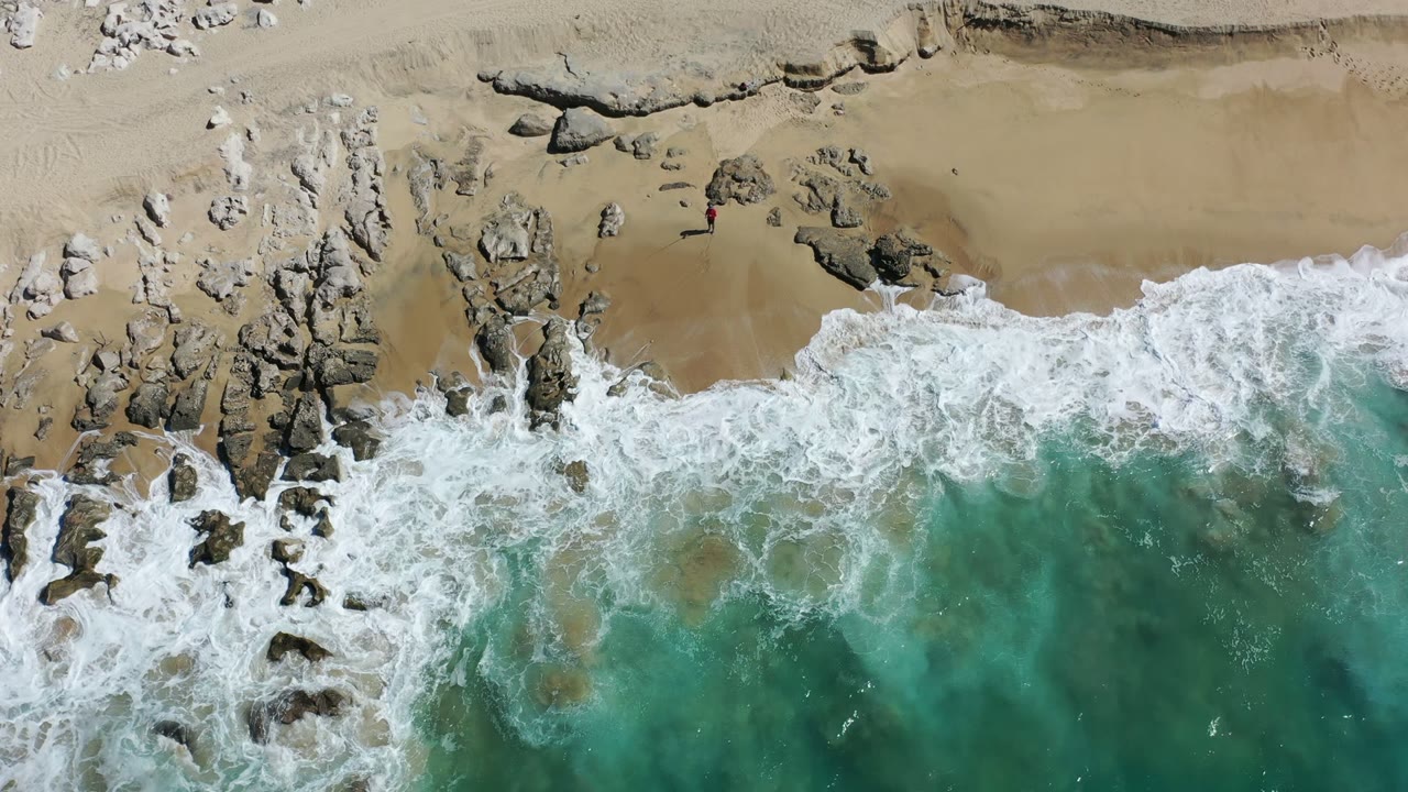 Los Cabos shoreline seen from above as white foam spreads farther across the rocks and tan beach, with no visible people.