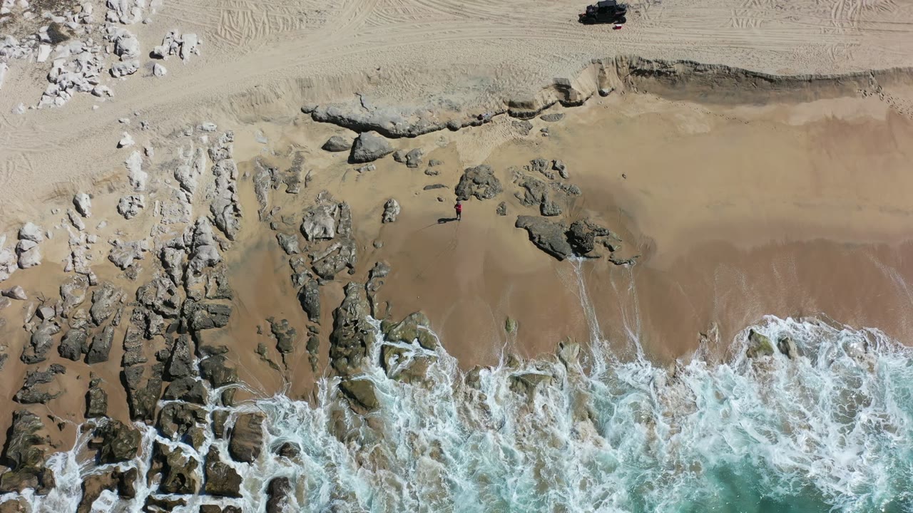 Overhead Los Cabos shoreline frame with foamy water spreading farther across the lower rocks, wet sand at center, dry tire-marked sand above, and no discernible people.