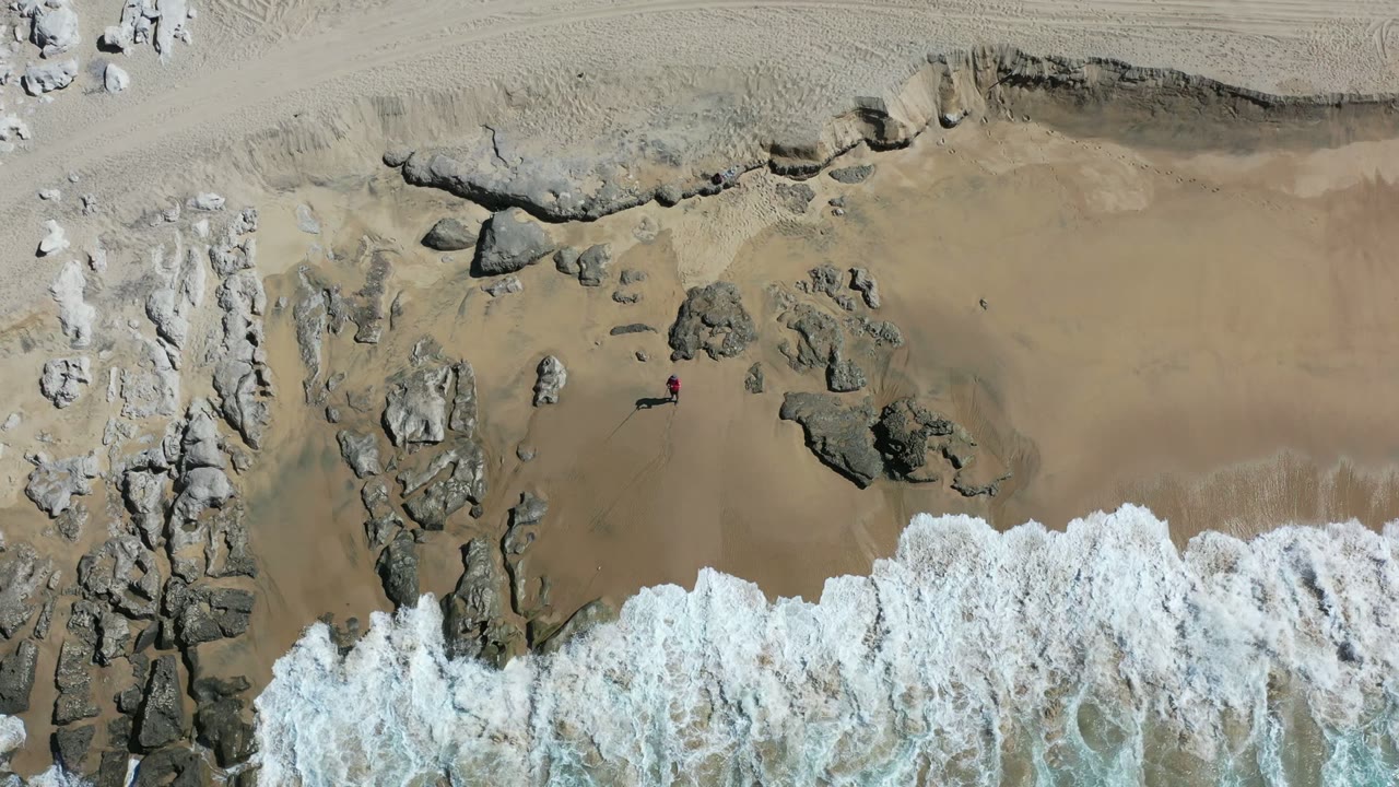 Los Cabos shoreline seen from overhead as a wider band of white foam spreads across the lower beach and around rock shelves, with no visible people.