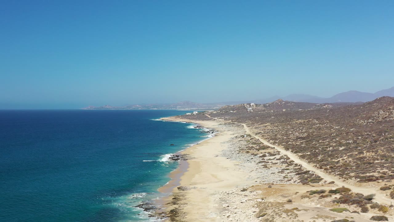 Wide aerial frame of the Los Cabos shoreline, with small white waves reaching the sand, darker rocks near the waterline, inland dirt paths, and no visible people.