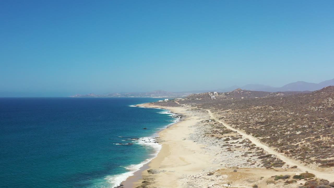 Aerial frame of the Los Cabos coast with deep blue ocean at left, white surf along a pale sandy beach, scrubby hills to the right, and no visible people.