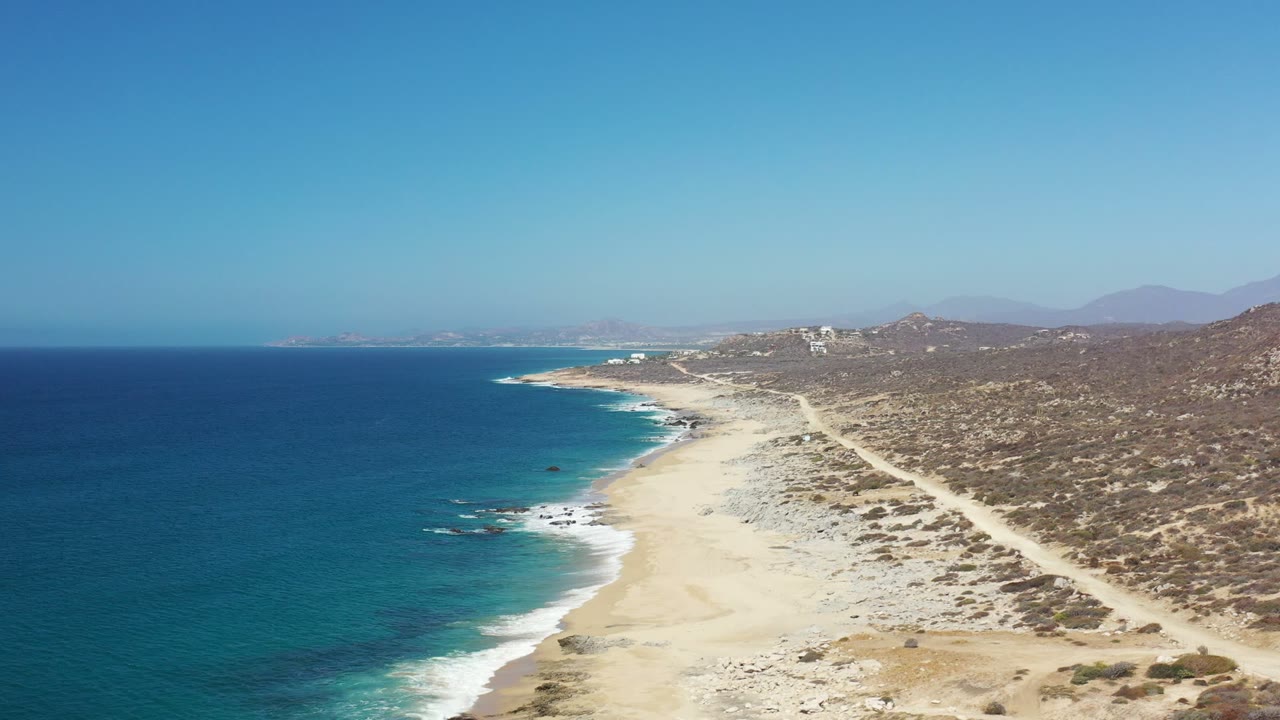 A second aerial view of the Los Cabos shoreline shows turquoise water near the surf line, a broad empty beach, dusty tracks inland, and no visible people.