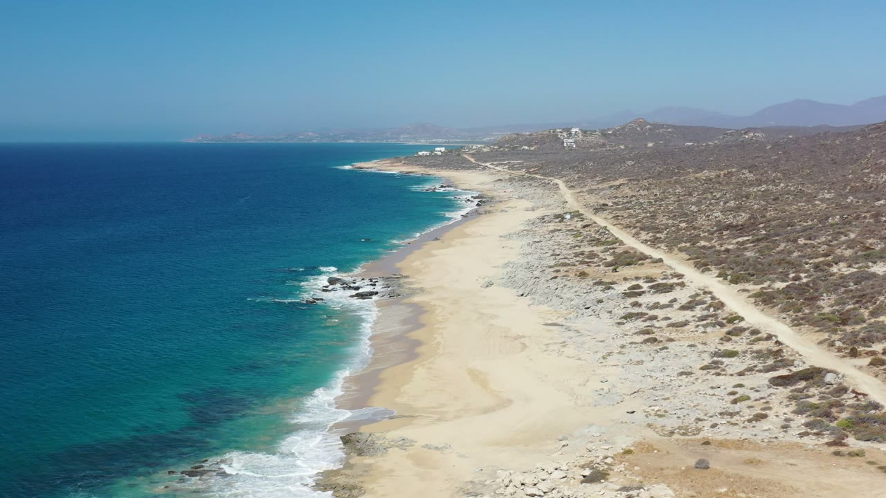 Los Cabos aerial frame with turquoise water closer to shore, thin white wave lines, a broad sandy beach, scrubby terrain, a diagonal dirt road, and no visible people.