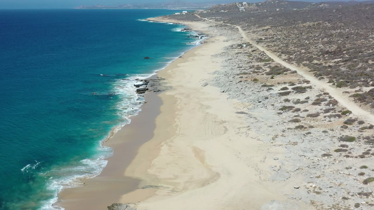 Aerial view in Los Cabos, Mexico, with turquoise water on the left, pale sand below, scrubby dunes to the right, and no visible people.