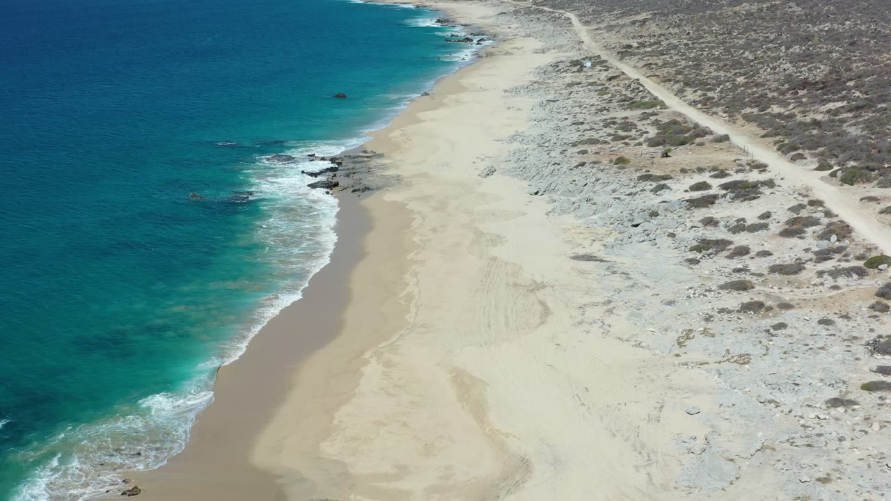 High angled view of the Los Cabos coast where blue water, a pale beach, scattered dark rocks, scrub dunes, and a dirt road fill the frame with no visible people.