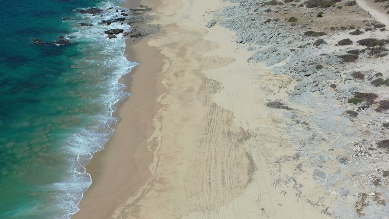 A Los Cabos shoreline frame shows foam sliding diagonally along wet sand, with rocky pale ground and scattered scrub filling the right side, no visible people.