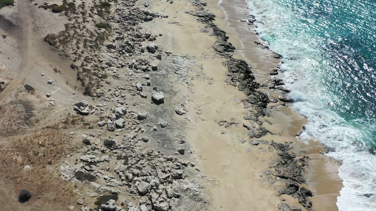 Overhead aerial view of a Los Cabos shoreline with rocky sand on the left, blue water on the right, white surf at the edge, and no visible people.