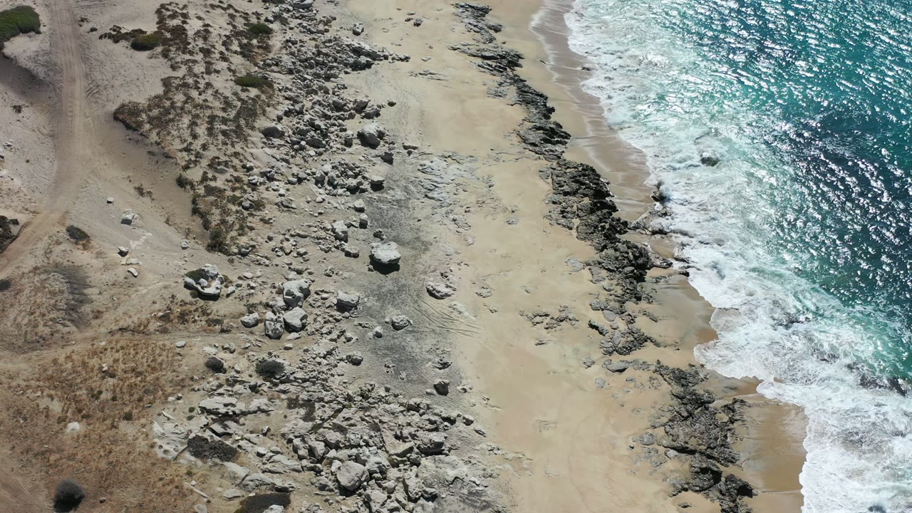 Overhead frame of the Los Cabos coast where white foam reaches a narrow sandy strip beside dark rocks and scrub-covered beach, with no visible people.