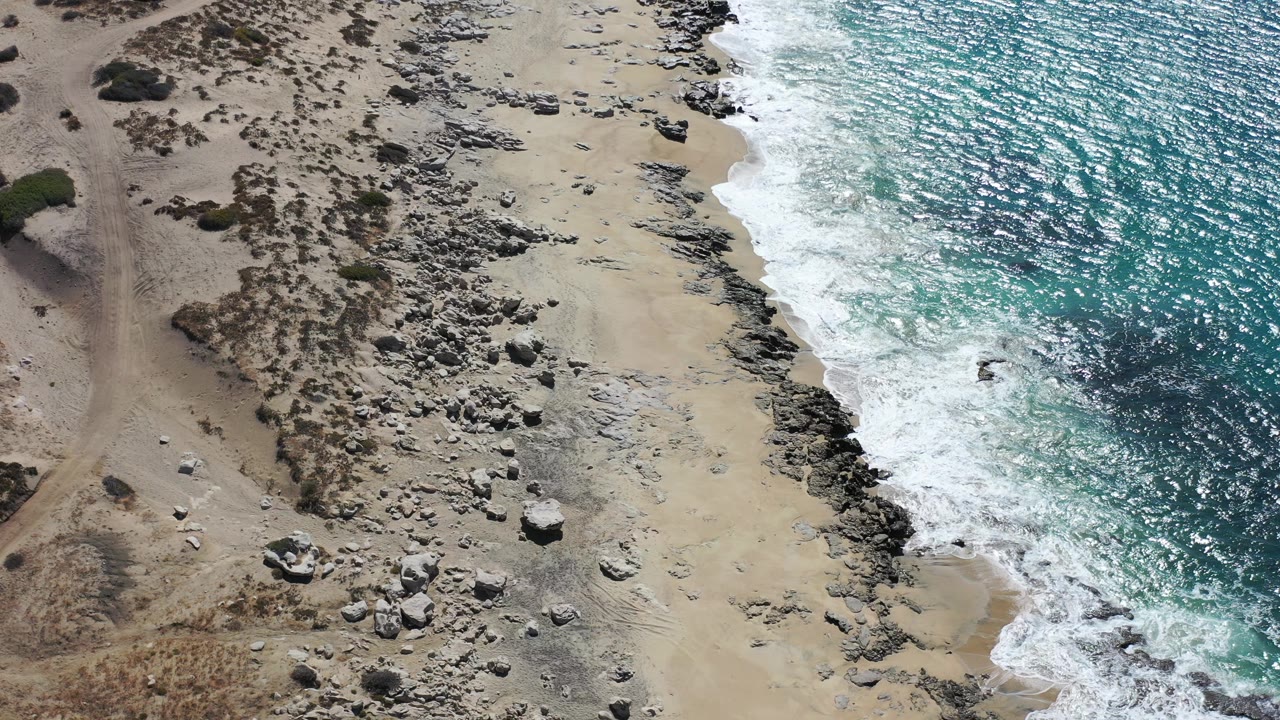 Aerial view of a Los Cabos beach with turquoise water breaking along dark rocks, pale sand, dry scrub, and no visible people.