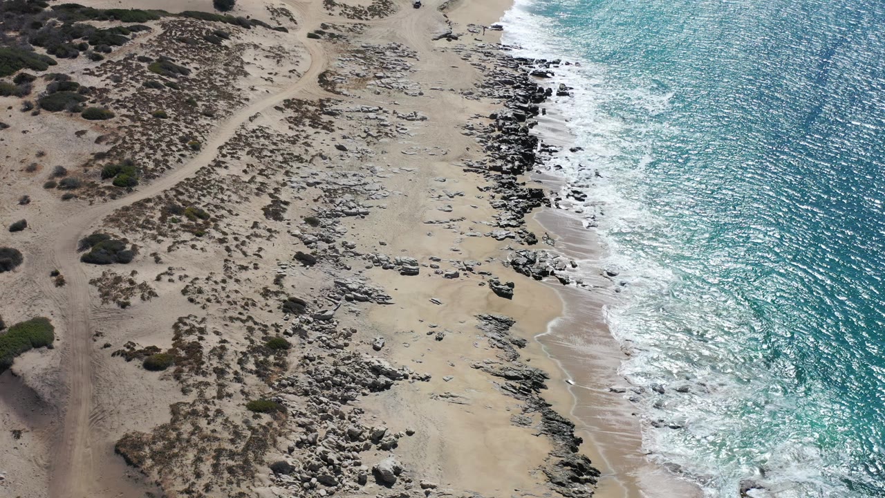 Aerial frame of Los Cabos shoreline where white water reaches the darker rock band beside open sand, with no visible people.