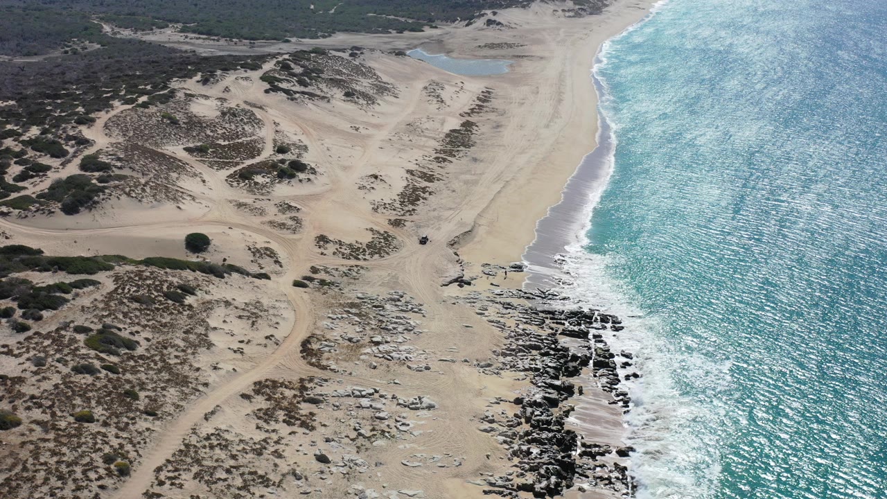 Opening aerial frame over a Los Cabos sandy beach with scrubby dunes inland, rock shelves near the shore, turquoise water at right, and no visible people.