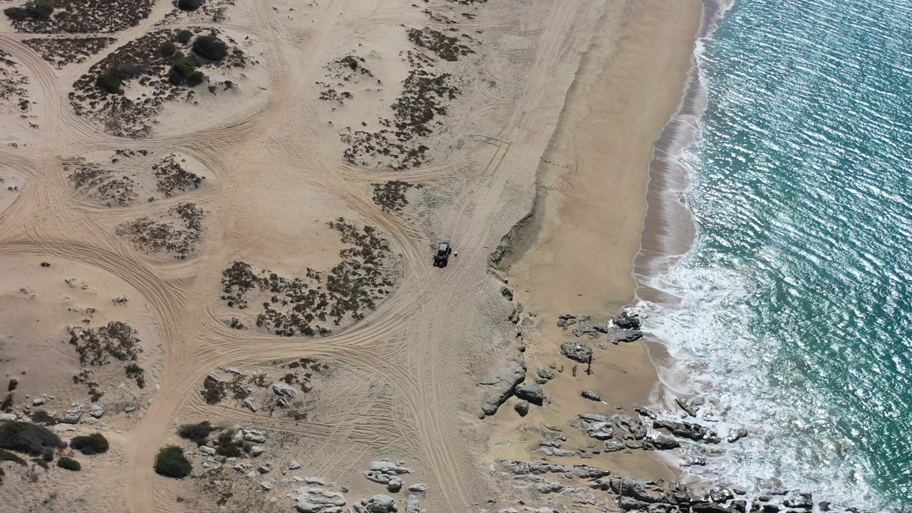 Overhead beach with dunes, tire tracks, rocks, and surf at frame start.