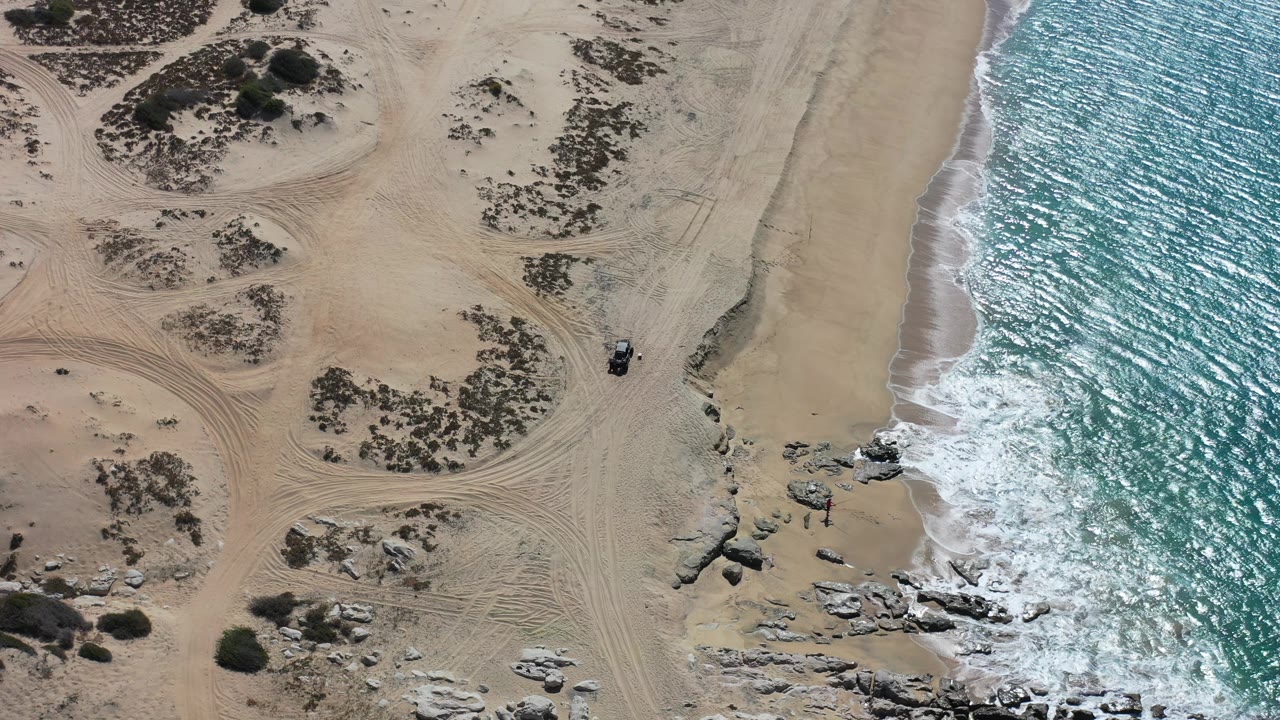 Aerial view over Los Cabos sand where tire tracks curve around low vegetation, a dark vehicle-like object sits near the beach edge, and white surf washes along the right side with no visible people.
