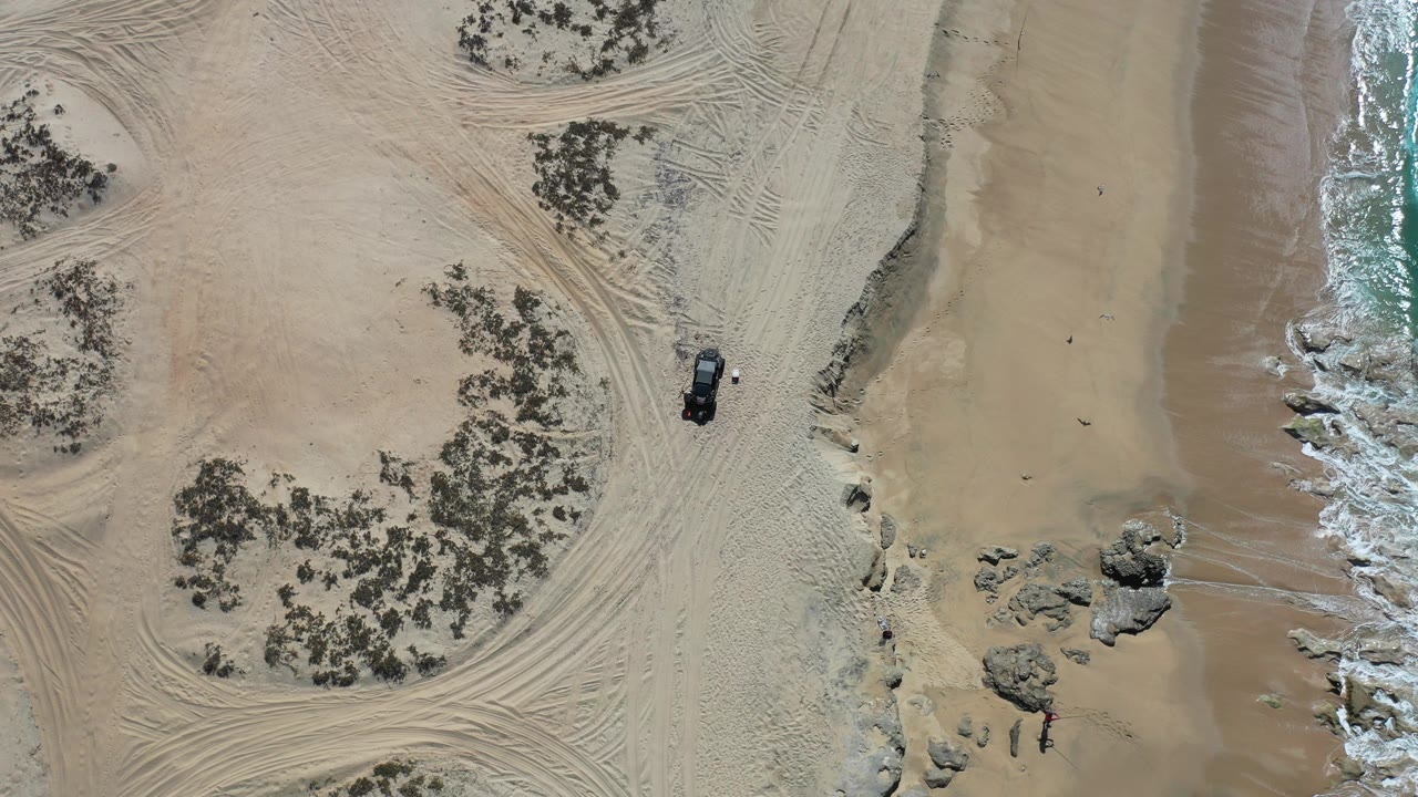 Top-down view over a Los Cabos beach with tire-marked sand, scattered scrub, shoreline rocks, white surf at the right edge, a black vehicle near center-right, and no visible people.