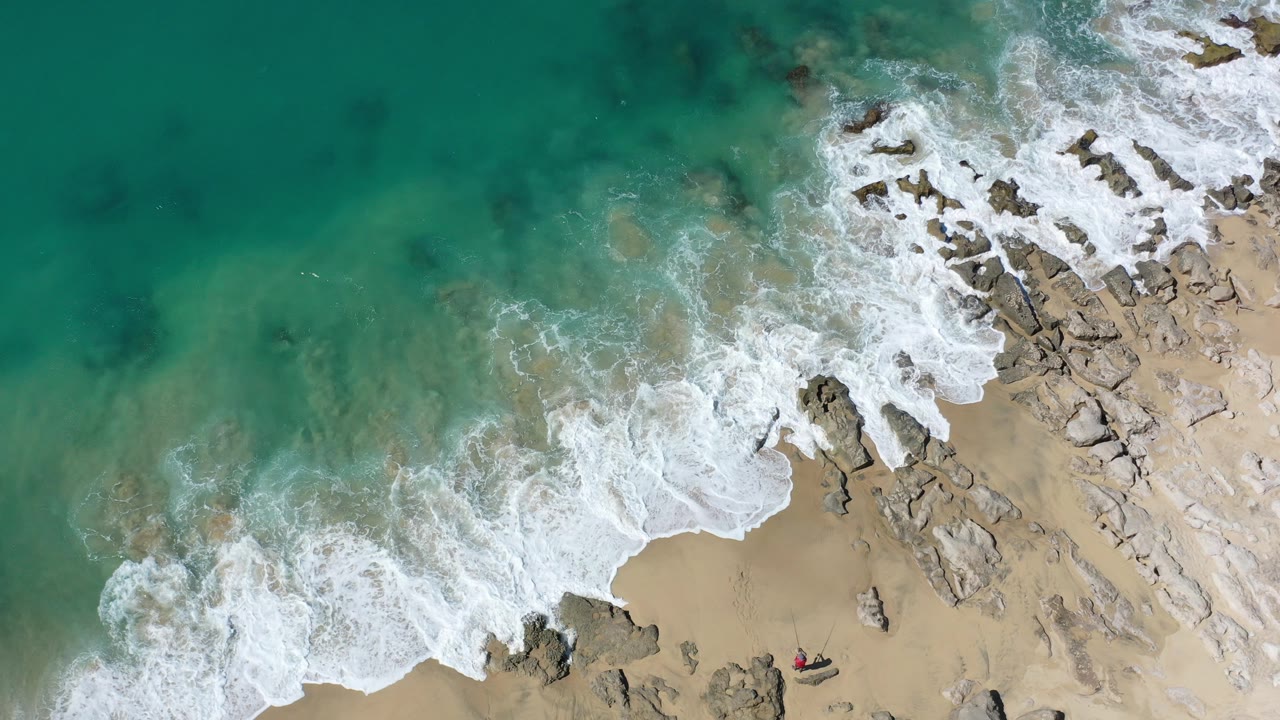 Overhead Los Cabos beach frame with turquoise water meeting pale sand and rock shelves, white surf spread across the lower left and right, and no visible people.