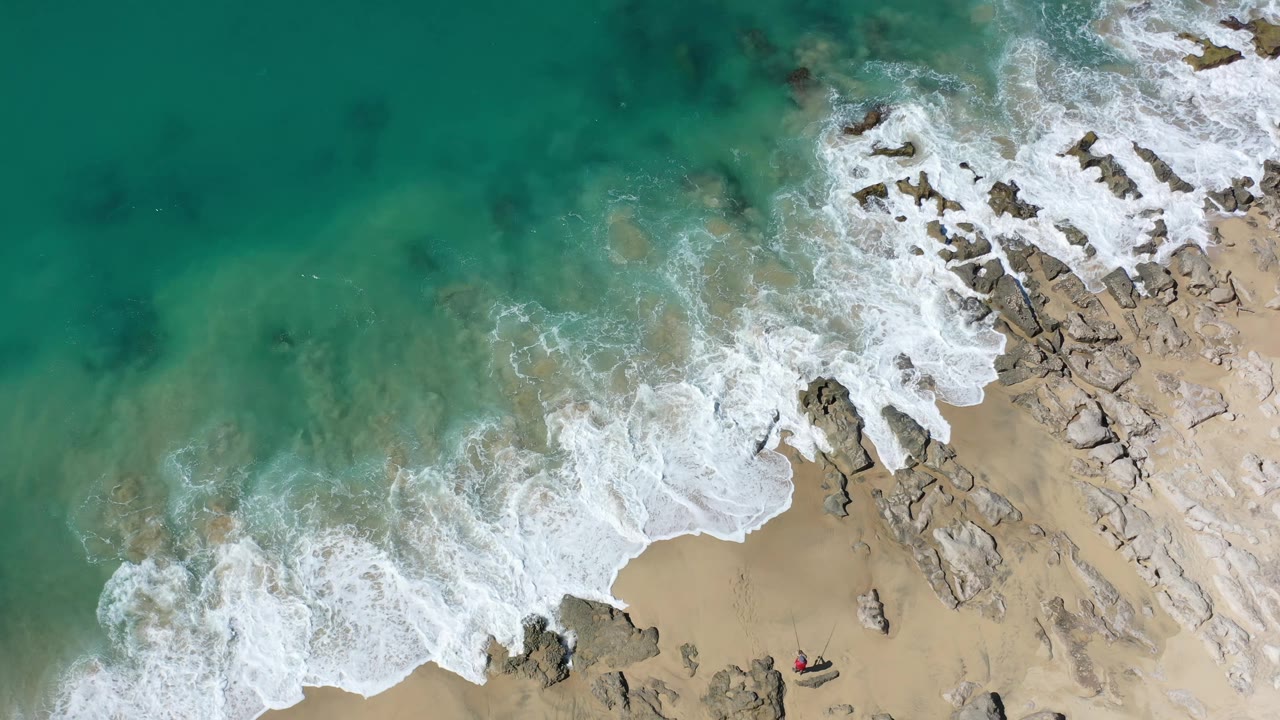 Aerial Los Cabos shoreline view where green water covers shallow rocks and white foam curls across the sand, with no visible people.