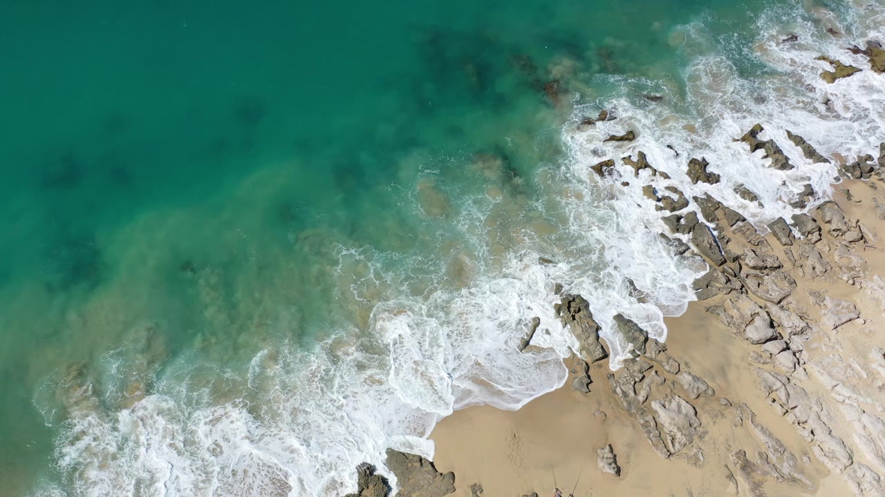 Top-down shoreline with surf foam, rocks, and clear green ocean.